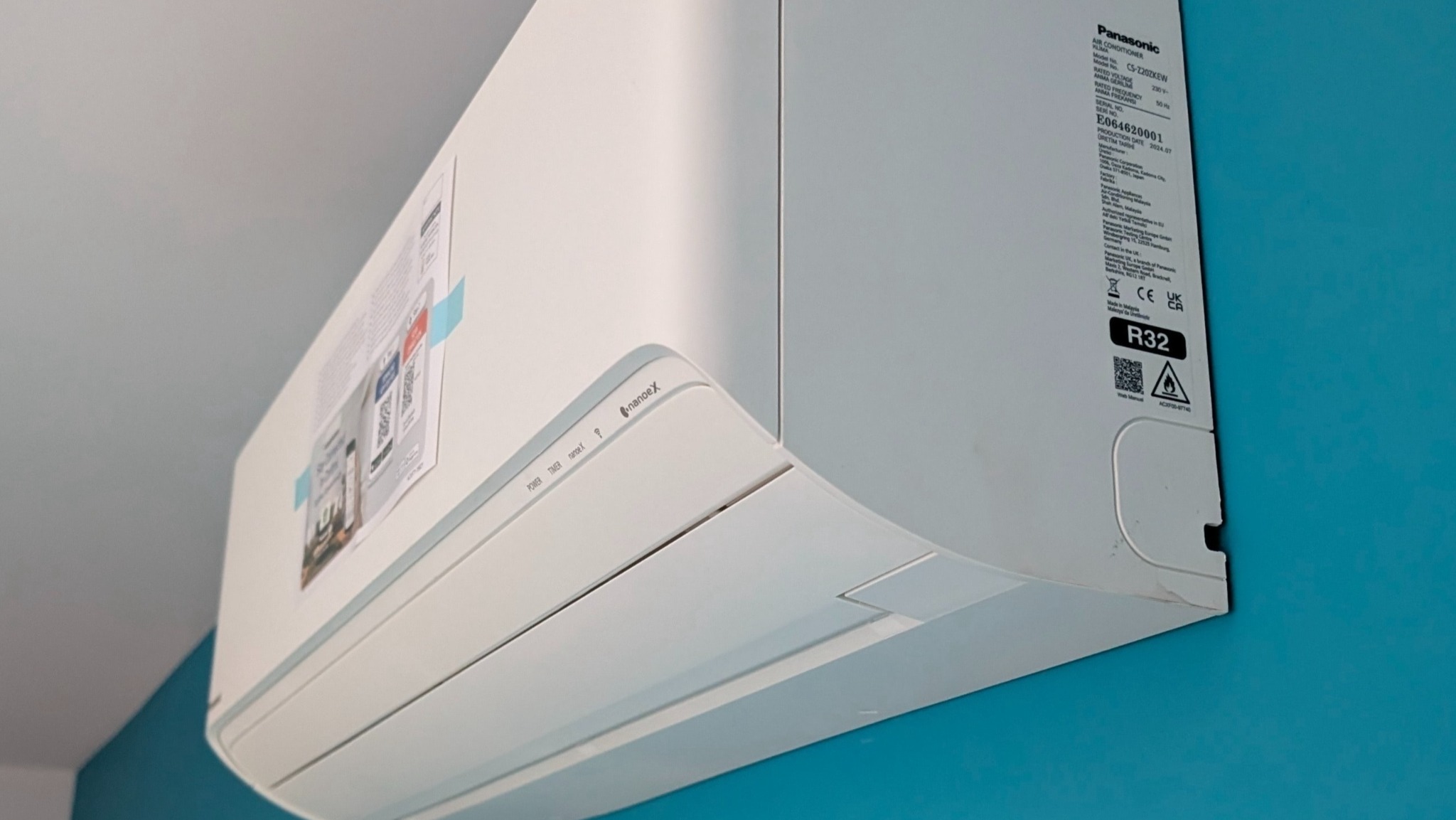 Biała klimatyzacja Panasonic na błękitnej ścianie, widok z dołu. Widoczne oznaczenia modelu i certyfikaty. Naklejka informacyjna na urządzeniu.