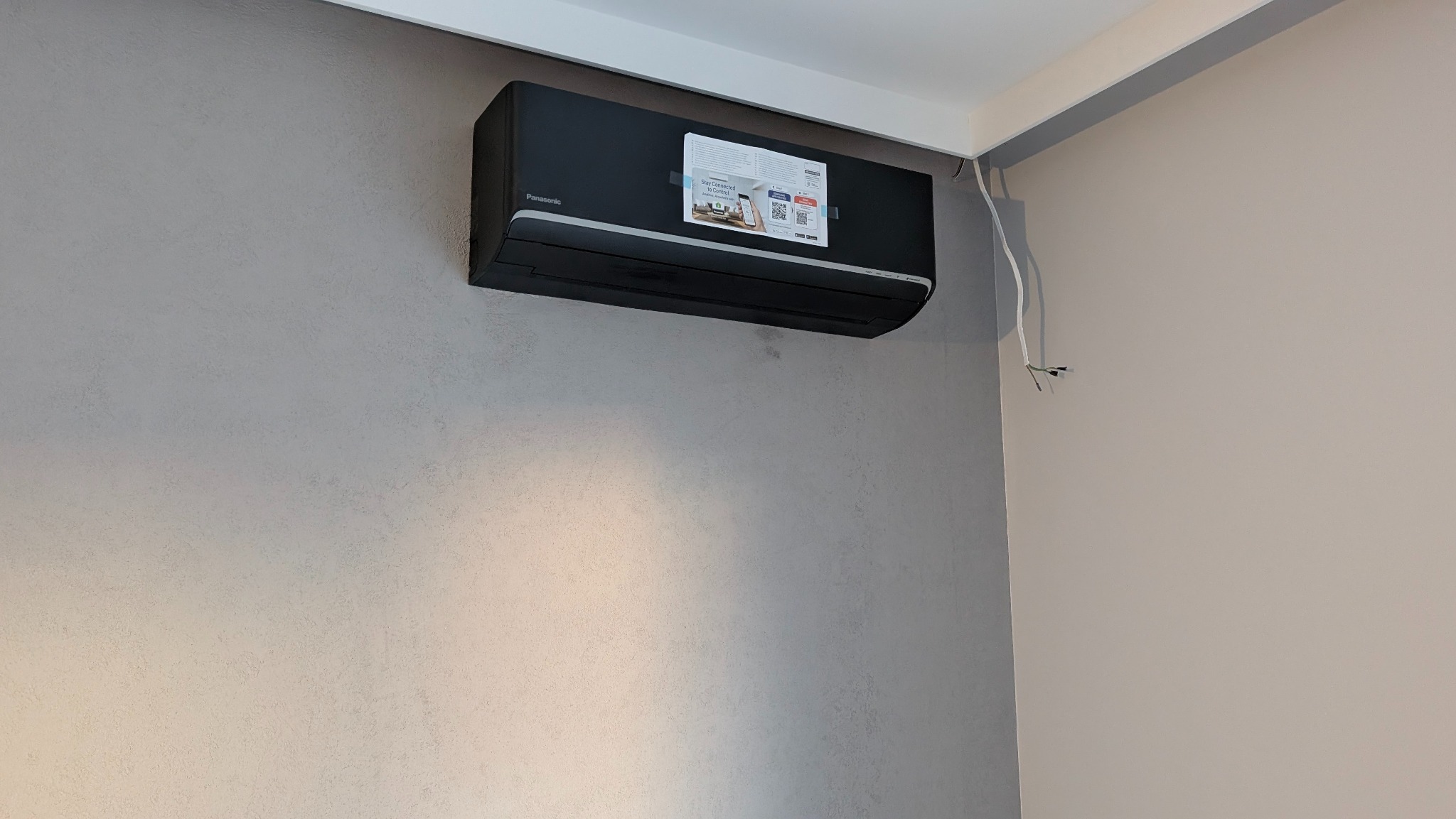 Czarny klimatyzator Panasonic zamontowany na szarej ścianie, widoczne przewody elektryczne. Minimalistyczny design, nowoczesne wnętrze.