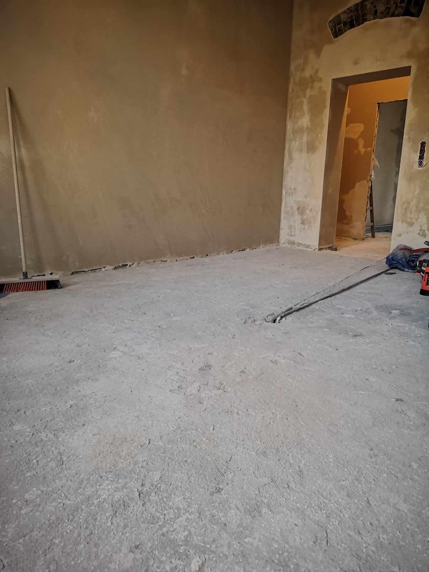 Pokój w trakcie remontu: ściany pokryte świeżym tynkiem, surowy beton na podłodze, widoczne narzędzia i szczotka przy ścianie, przejście do kolejnego pomieszczenia z drabiną.