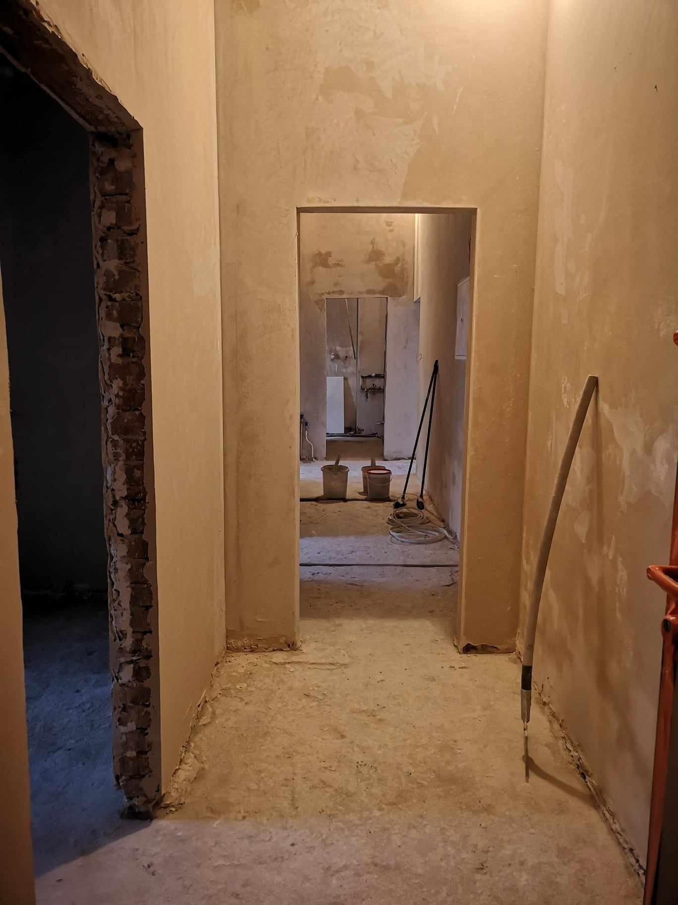 Korytarz w trakcie remontu, ściany przygotowane pod wykończenie, widoczne ślady po skrobaniu, surowy beton na podłodze, dwa otwory drzwiowe, jeden z widoczną cegłą. W tle wiaderka i sprzęt malarski.