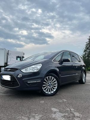 Ciemnoszary Ford S-Max zaparkowany w Sochaczewie, widok z przodu i boku, z widocznymi aluminiowymi felgami i odblaskami na karoserii. W tle widoczny fragment ciężarówki i pochmurne niebo.