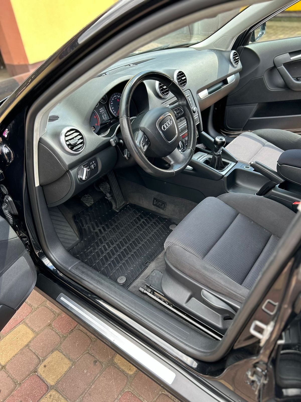 Wnętrze czarnego samochodu Audi A3 z widocznymi szarymi fotelami, gumowymi dywanikami i deską rozdzielczą z kierownicą.