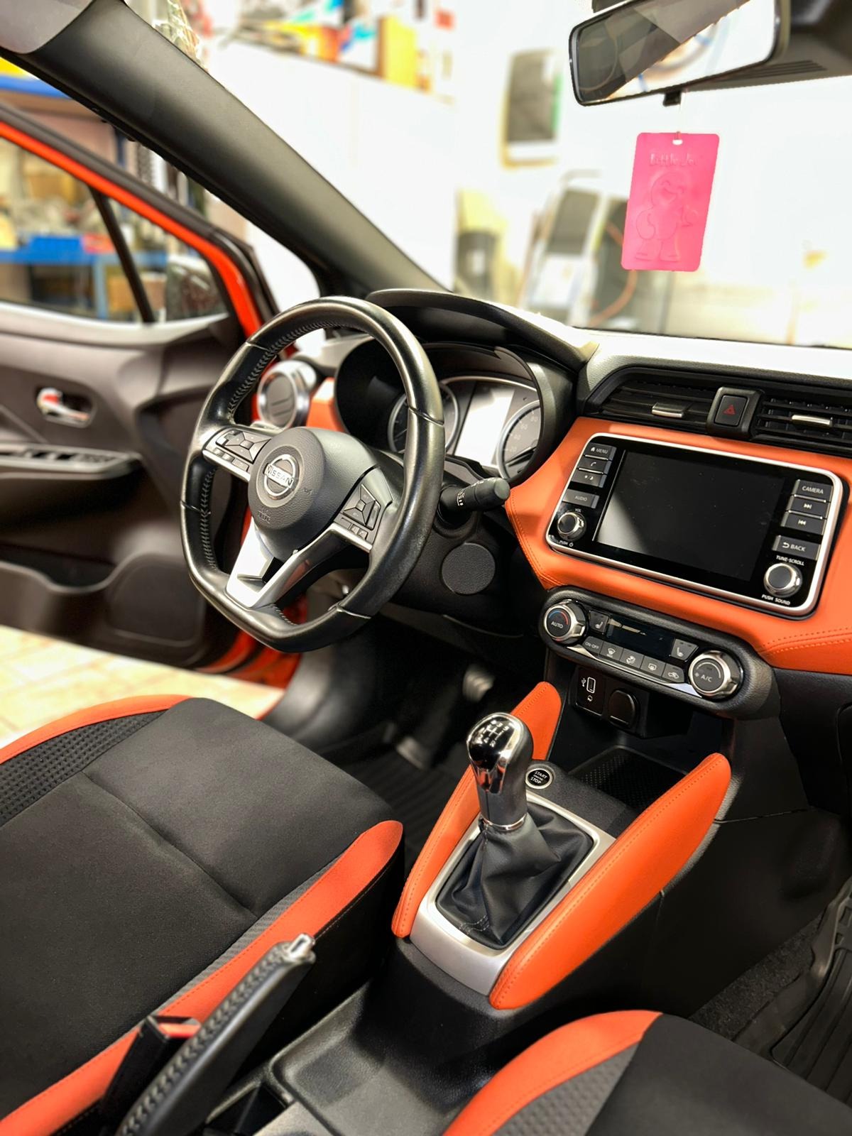 Wnętrze samochodu Nissan Micra z czarno-pomarańczową tapicerką, widoczna kierownica, deska rozdzielcza i dźwignia zmiany biegów. Na lusterku zawieszka zapachowa.