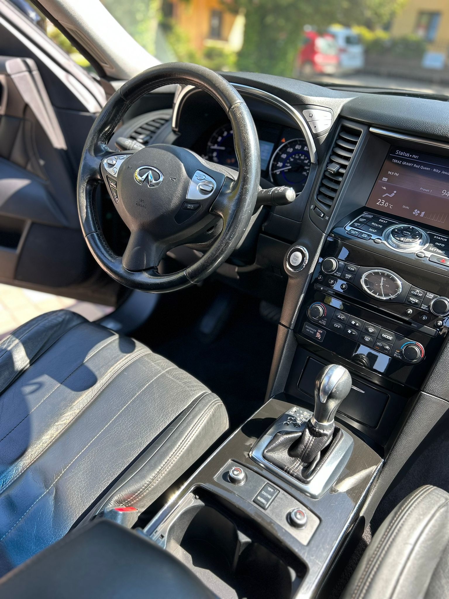 Wnętrze samochodu marki Infiniti z widoczną deską rozdzielczą, kierownicą, ekranem dotykowym i dźwignią zmiany biegów. Na skórzanych siedzeniach widoczne refleksy słoneczne.