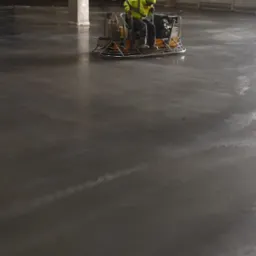 Pracownik w odblaskowej kamizelce, wygładza wylewkę betonową za pomocą zacieraczki mechanicznej na dużej powierzchni przemysłowej.