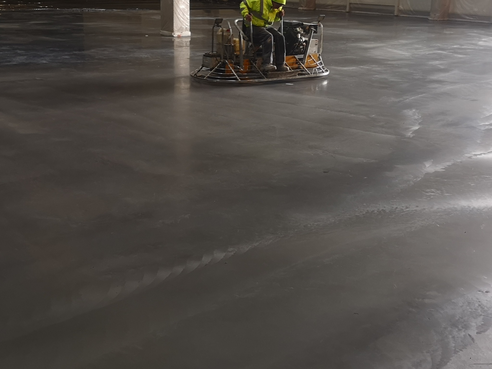 Pracownik w odblaskowej kamizelce, wygładza wylewkę betonową za pomocą zacieraczki mechanicznej na dużej powierzchni przemysłowej.