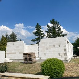 Bahniuk-Bud Bahniuk Anton - Wznoszone ściany parterowego budynku z białych bloczków, widoczne palety z materiałem budowlanym, błękitne niebo z białymi chmurami w tle.