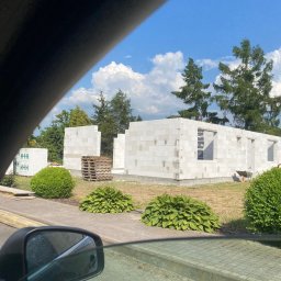 Bahniuk-Bud Bahniuk Anton - Wznoszone ściany parterowego budynku z białych bloczków, widok zza szyby samochodu, palety z materiałem budowlanym w tle, zielona trawa i krzewy.