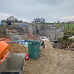 Bahniuk-Bud Bahniuk Anton - Budowa domu murowanego w trakcie: wznoszone ściany z cegły i bloczków, widoczny murarz, betoniarka, taczka z zaprawą, teren budowy z widokiem na domy jednorodzinne w tle.