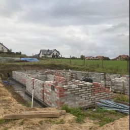 Bahniuk-Bud Bahniuk Anton - Wznoszenie ścian fundamentowych z bloczków betonowych i cegieł klinkierowych na terenie budowy domu jednorodzinnego, widoczne izolowane fundamenty i przygotowane zbrojenie, w tle zabudowa...