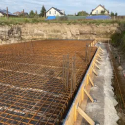 Zbrojenie fundamentu w szalunku drewnianym, widok z góry na siatkę z prętów stalowych, w tle wykop ziemny i zabudowa jednorodzinna.