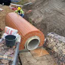 Budowa przepustu z cegły klinkierowej z rurą betonową wewnątrz, zabezpieczonego gabionami z kamienia, pracownik w kasku kontroluje wykonanie na placu budowy.