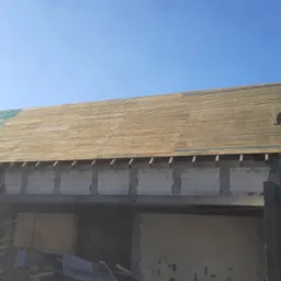 Wznoszony dom murowany z częściowo ułożoną drewnianą konstrukcją dachu, widoczne bloczki betonowe i elementy więźby dachowej, na tle błękitnego nieba.