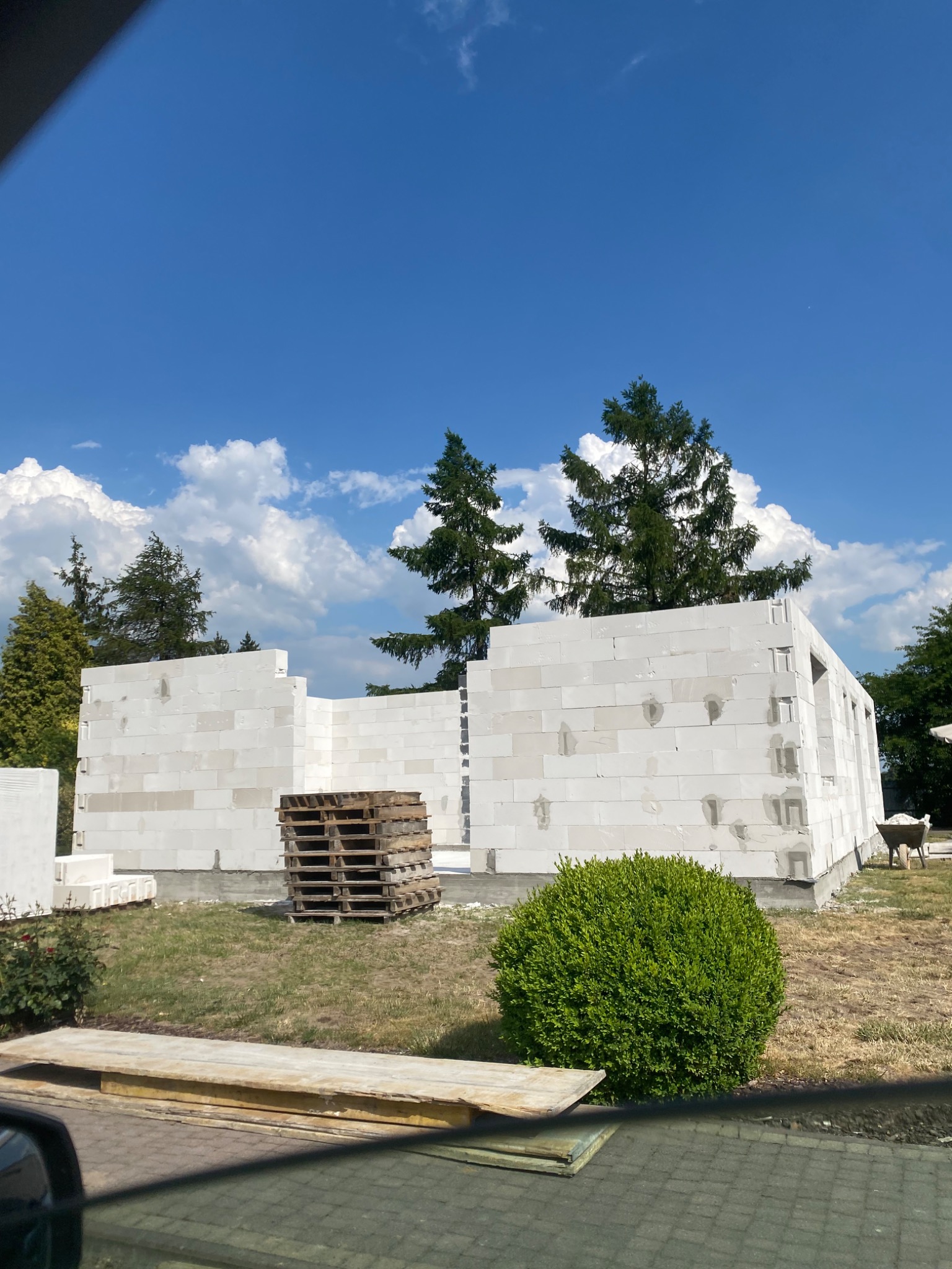 Wznoszone ściany parterowego budynku z białych bloczków, widoczne palety z materiałem budowlanym, błękitne niebo z białymi chmurami w tle.