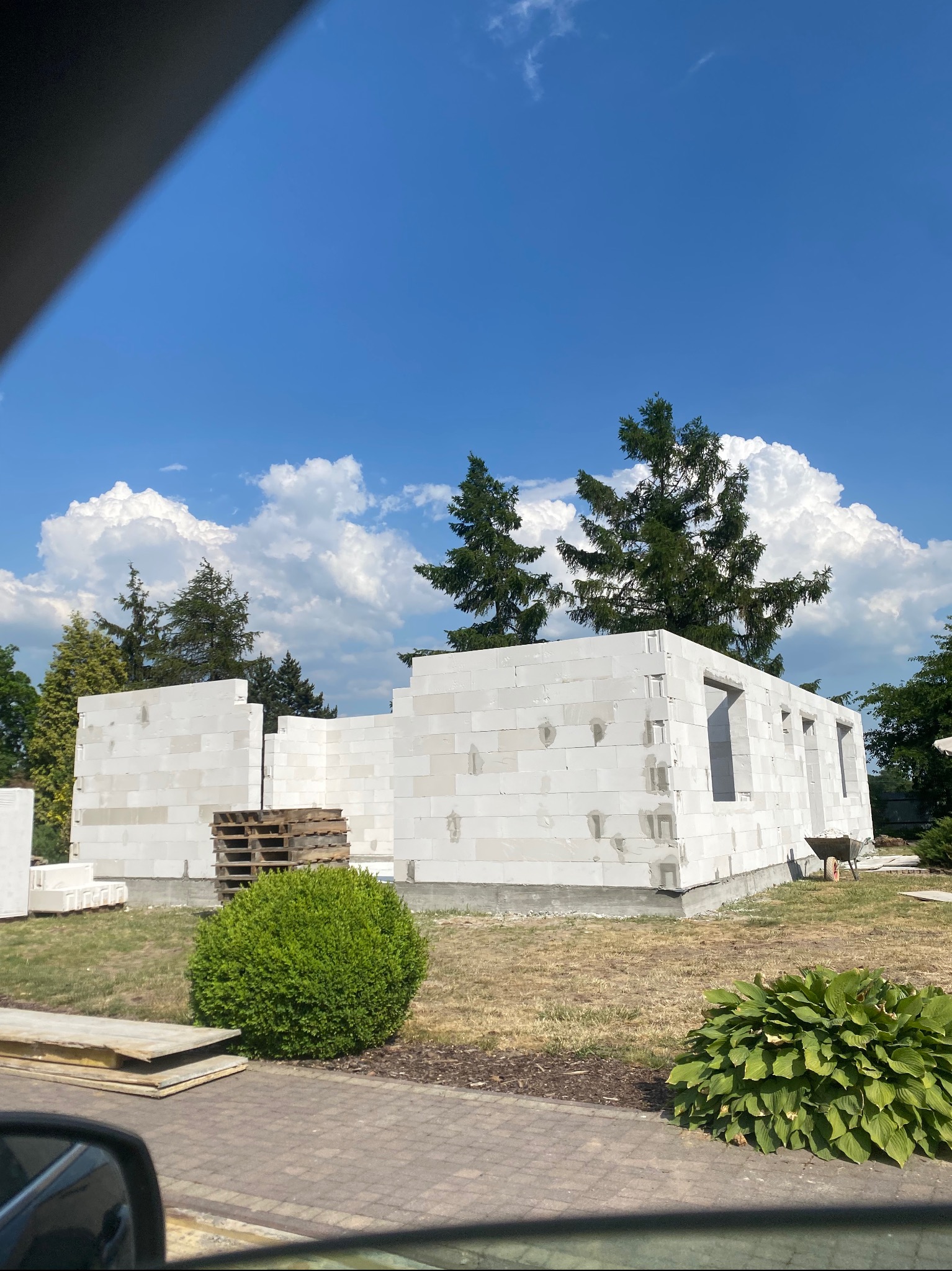 Budowa domu z białych bloczków silikatowych w słoneczny dzień, widoczne palety z materiałem budowlanym i taczka, w tle drzewa i błękitne niebo z chmurami.