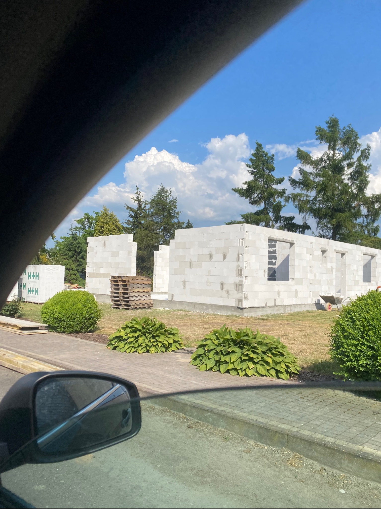 Wznoszone ściany parterowego budynku z białych bloczków, widok zza szyby samochodu, palety z materiałem budowlanym w tle, zielona trawa i krzewy.