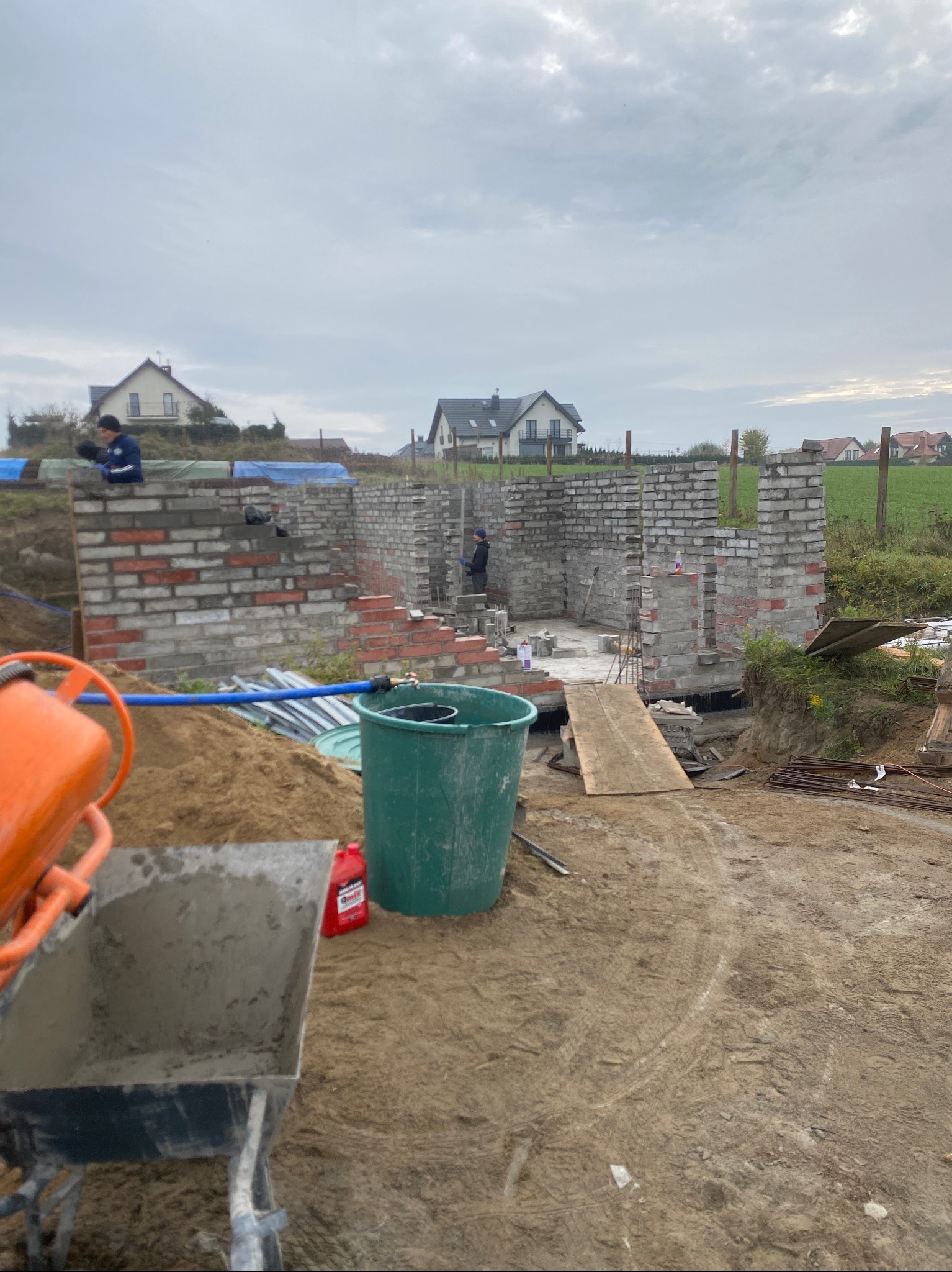 Budowa domu murowanego w trakcie: wznoszone ściany z cegły i bloczków, widoczny murarz, betoniarka, taczka z zaprawą, teren budowy z widokiem na domy jednorodzinne w tle.