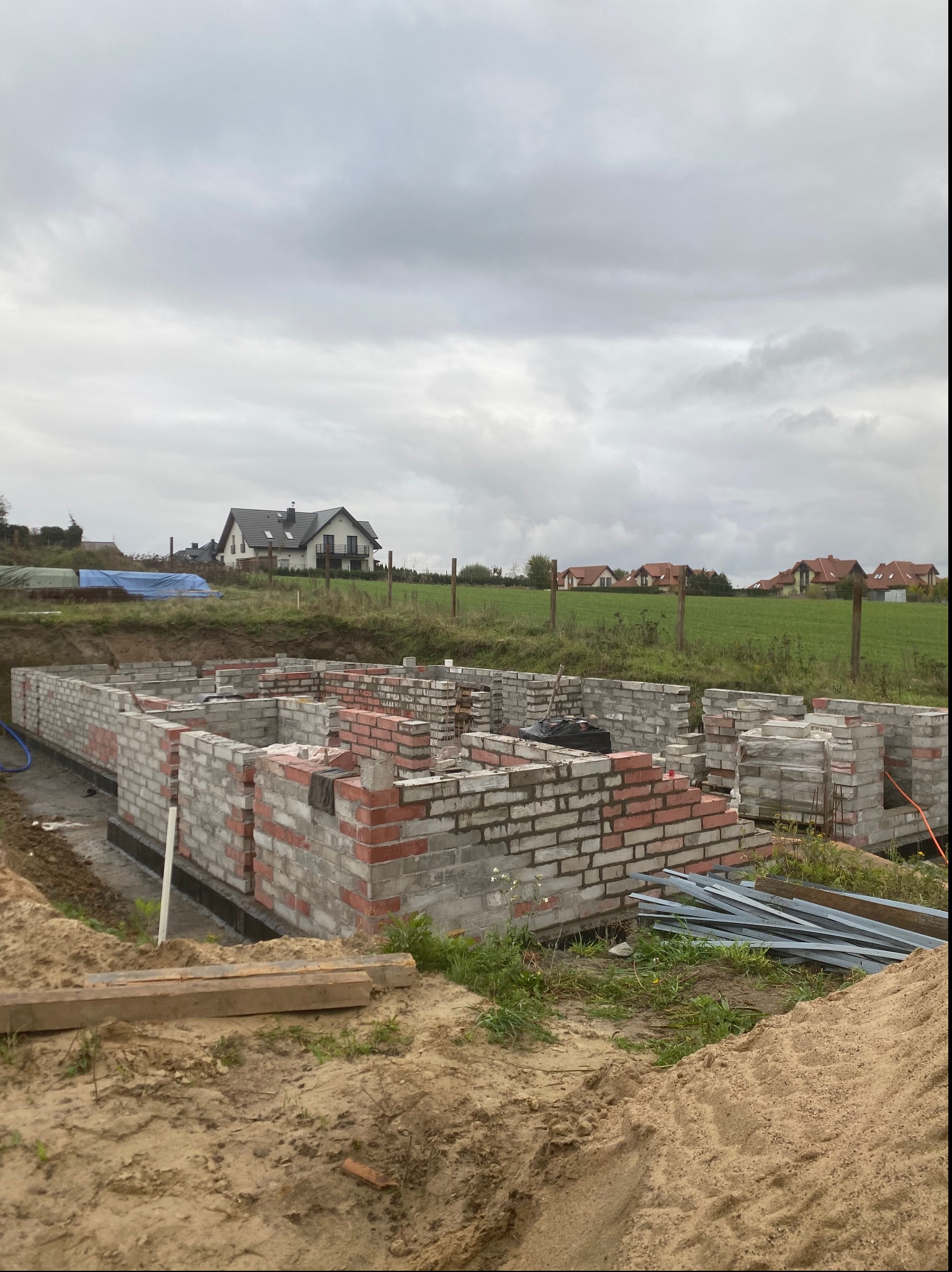 Budowa domu jednorodzinnego: wzniesione mury fundamentowe z czerwonej i szarej cegły silikatowej na tle zielonego krajobrazu i pochmurnego nieba.
