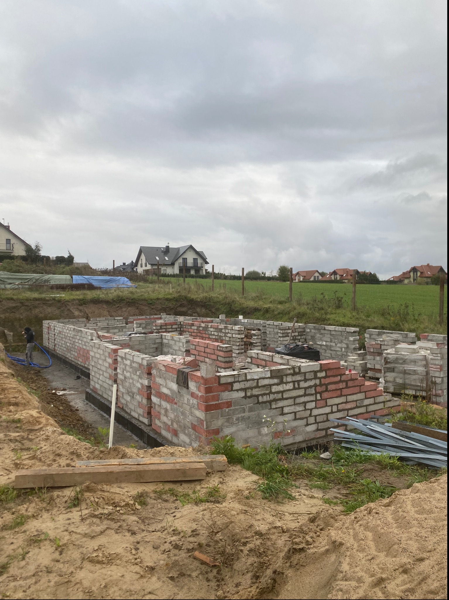 Wznoszenie ścian fundamentowych z bloczków betonowych i cegieł klinkierowych na terenie budowy domu jednorodzinnego, widoczne izolowane fundamenty i przygotowane zbrojenie, w tle zabudowa...