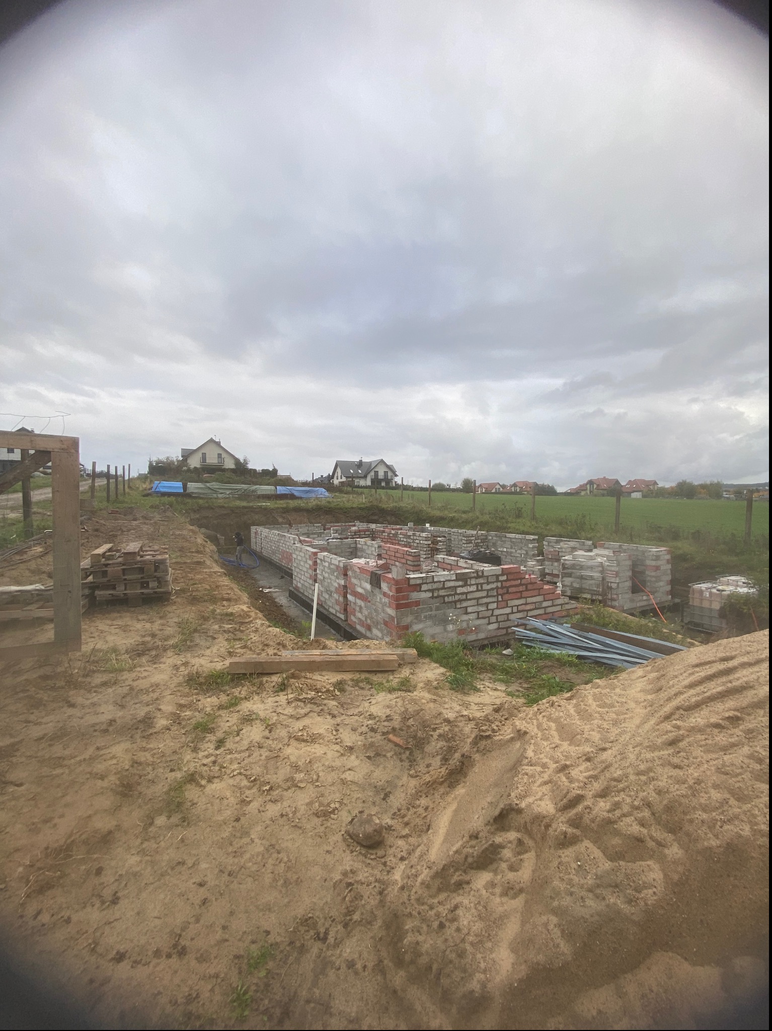 Wznoszone ściany fundamentowe z czerwonej cegły silikatowej na budowie domu jednorodzinnego, widok na okoliczne domy i pochmurne niebo.