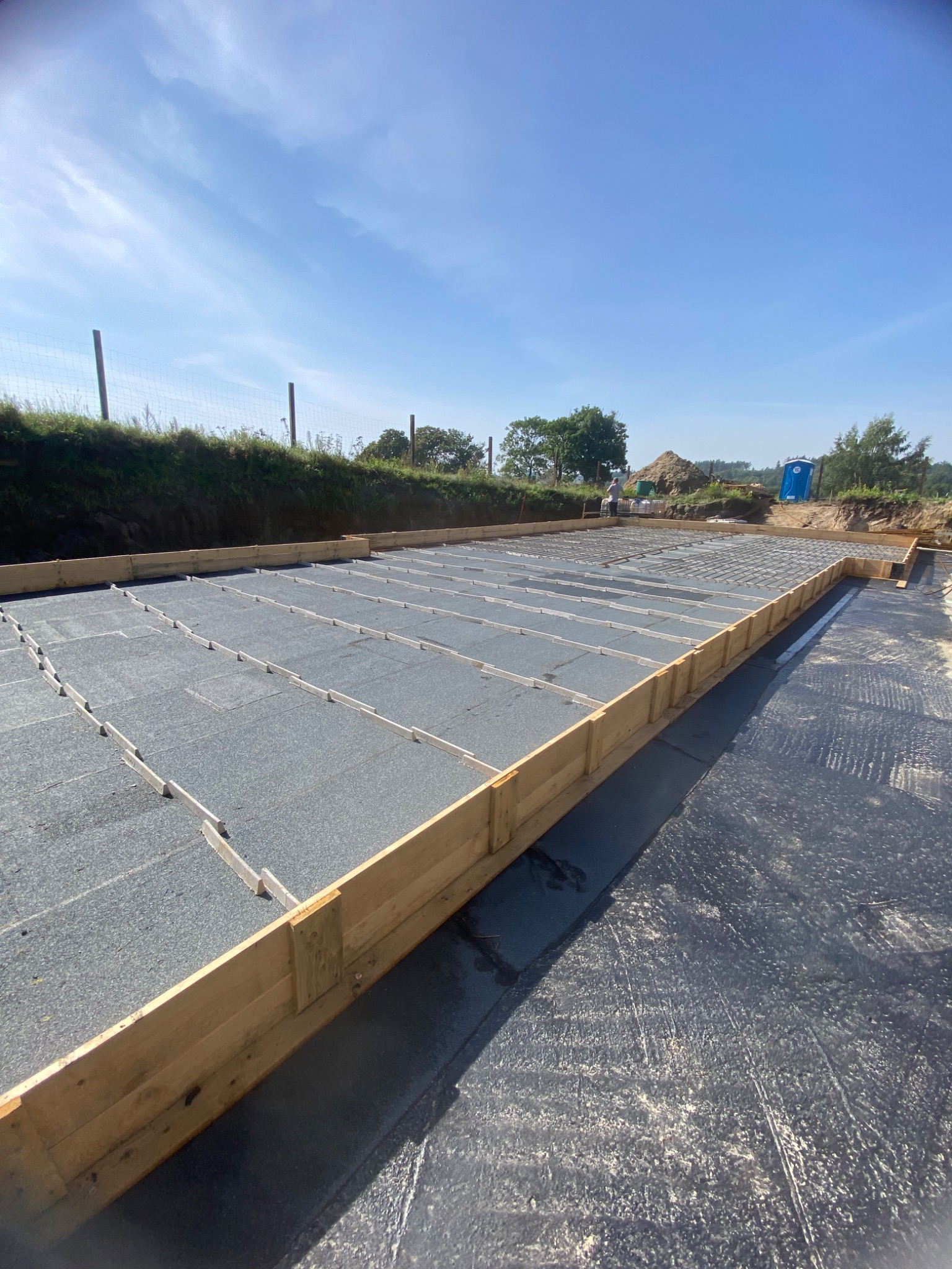 Przygotowany teren pod fundamenty z widoczną folią hydroizolacyjną, drewnianym szalunkiem i zbrojeniem siatką stalową, w tle widoczna ziemia, drzewa i niebieska toaleta przenośna.