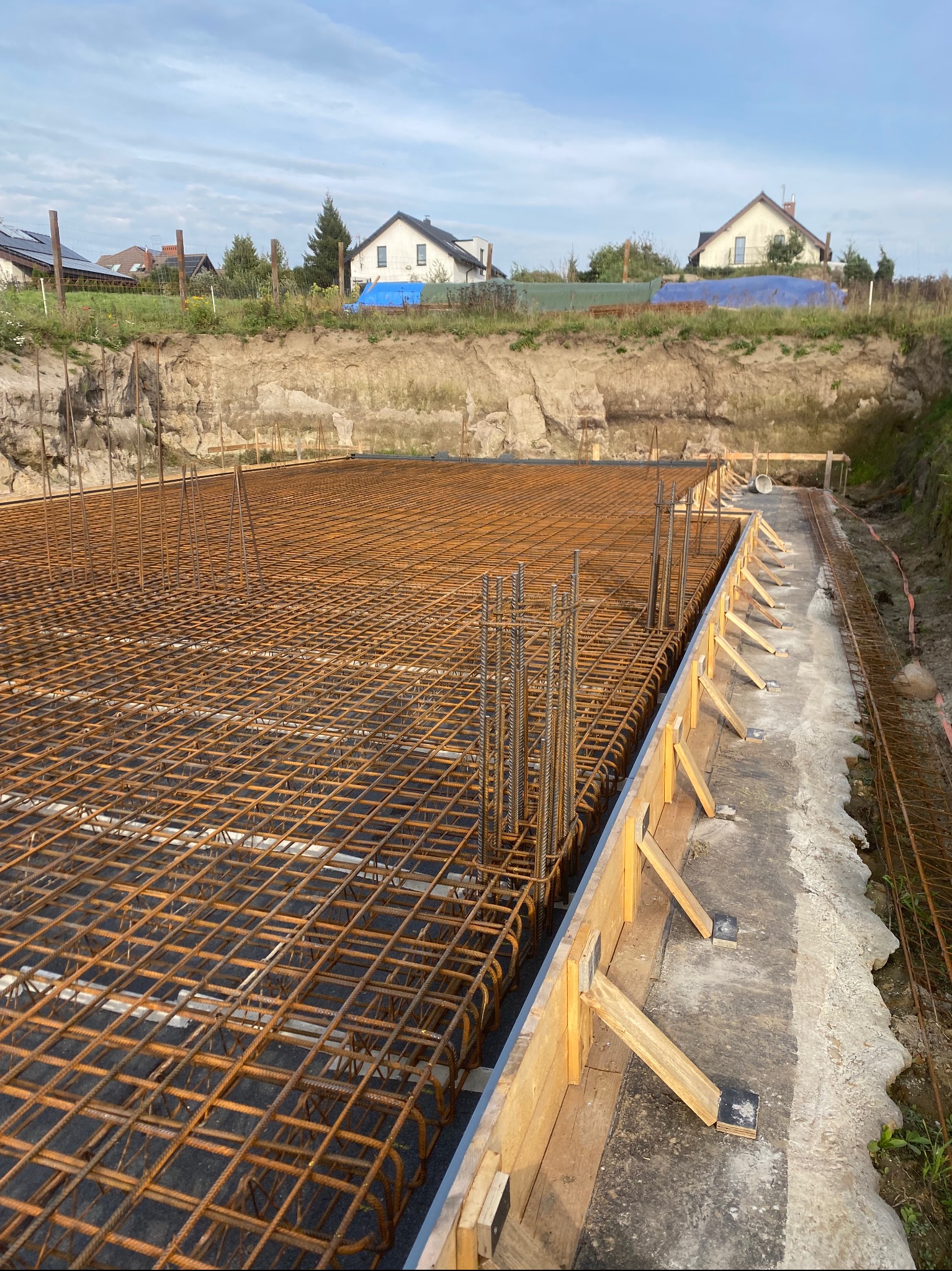Zbrojenie fundamentu w szalunku drewnianym, widok z góry na siatkę z prętów stalowych, w tle wykop ziemny i zabudowa jednorodzinna.