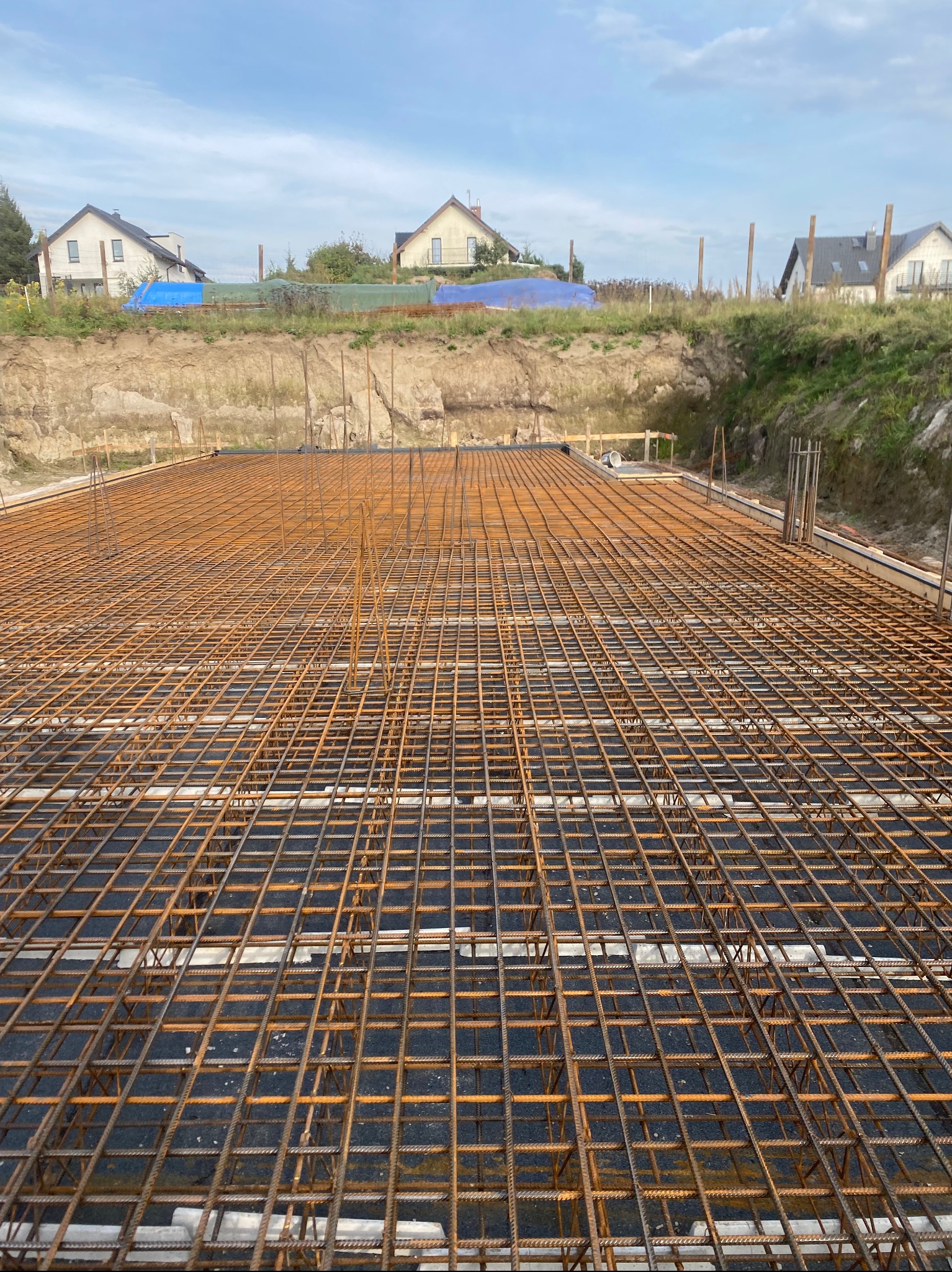 Zbrojenie fundamentu domu jednorodzinnego, widok z góry na siatkę z prętów zbrojeniowych, w tle wykop i zabudowa jednorodzinna.