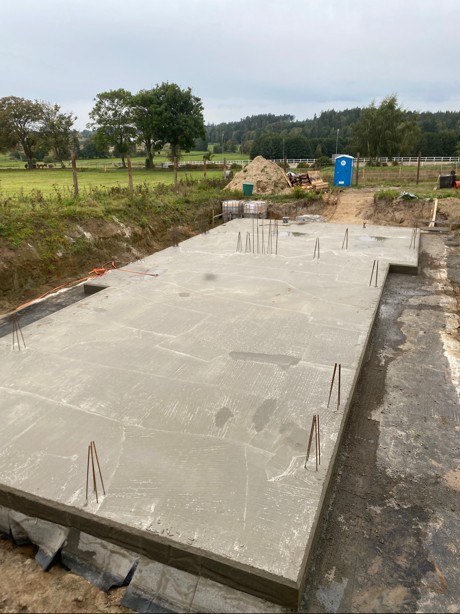 Wylany fundament płyty betonowej z wystającymi prętami zbrojeniowymi, widoczne zabezpieczenie przeciwwilgociowe, w tle teren budowy z pryzmą piasku i przenośną toaletą.