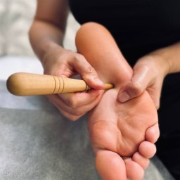Shchetinina Larysa - Masaż stóp drewnianym przyrządem na leżance w gabinecie. Dłoń terapeuty uciska punkt na stopie pacjenta. Bliskie ujęcie.