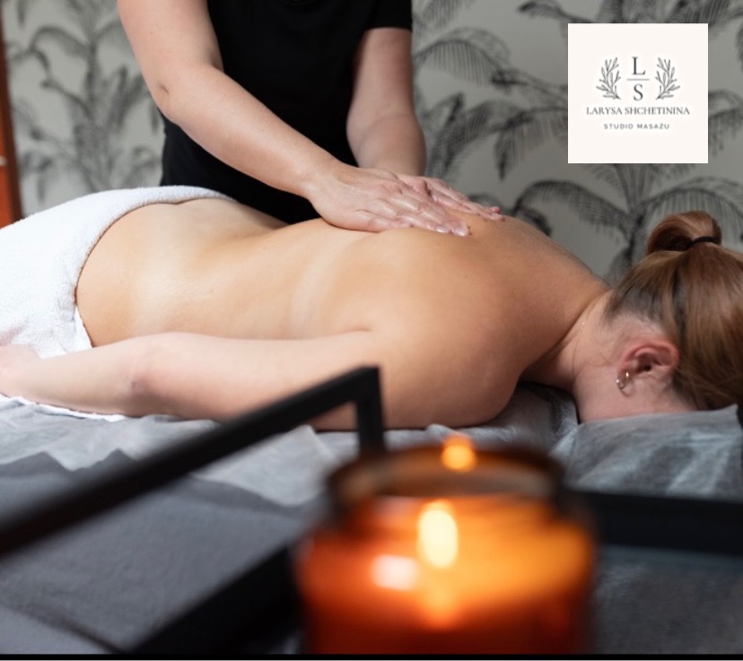 Kobieta podczas masażu pleców w salonie, widoczna zapalona świeca i logo salonu na ścianie.
