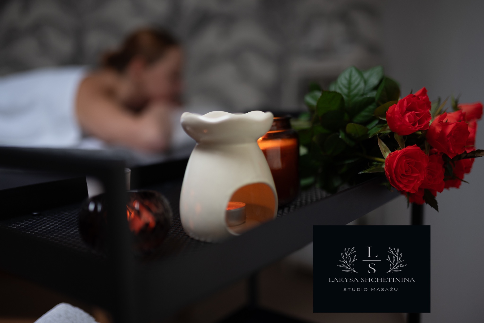 Kadr z gabinetu: rozmyta sylwetka osoby leżącej na stole do zabiegów w tle, aromaterapeutyczny podgrzewacz olejku z zapaloną świeczką, bukiet czerwonych róż i logo studia na pierwszym planie.
