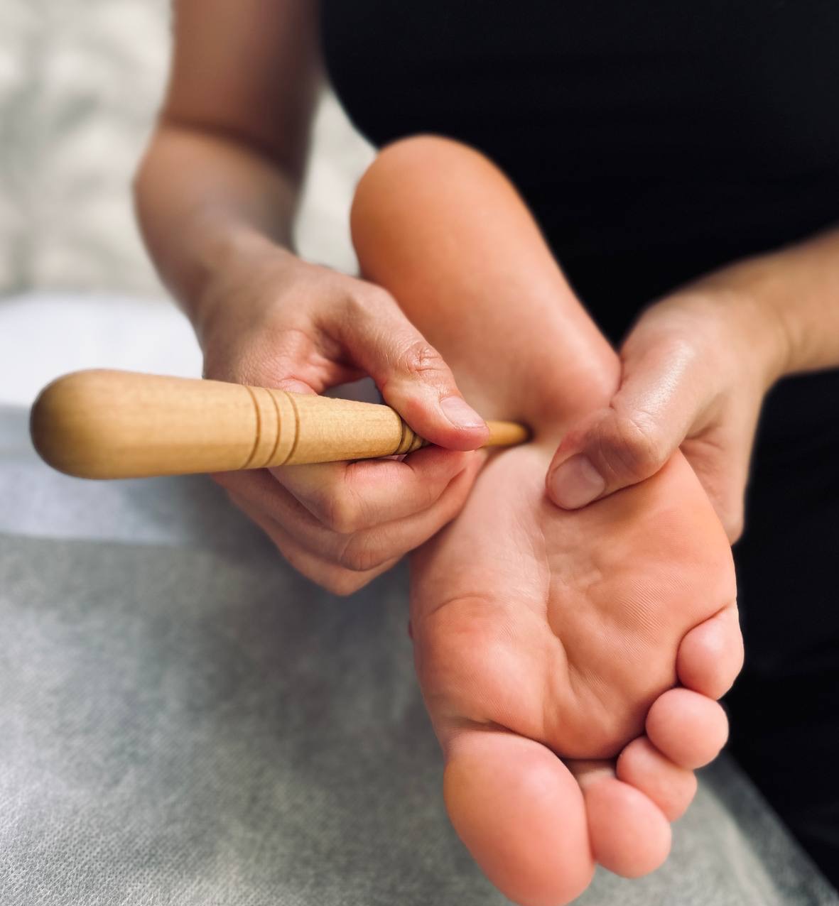Masaż stóp drewnianym przyrządem na leżance w gabinecie. Dłoń terapeuty uciska punkt na stopie pacjenta. Bliskie ujęcie.