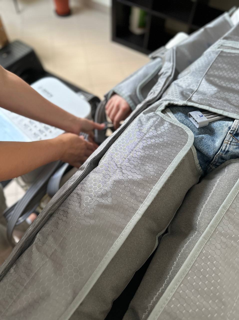 Osoba w kombinezonie do presoterapii, z widocznymi przewodami i dłońmi terapeuty dopinającymi pasy. Urządzenie obok. Wnętrze gabinetu.