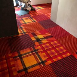 SALBUD Artur Salwach - Układanie wzorzystej, czerwono-pomarańczowej wykładziny podłogowej w kratę przez montera w pomieszczeniu z białymi ścianami i ciemnymi drzwiami.