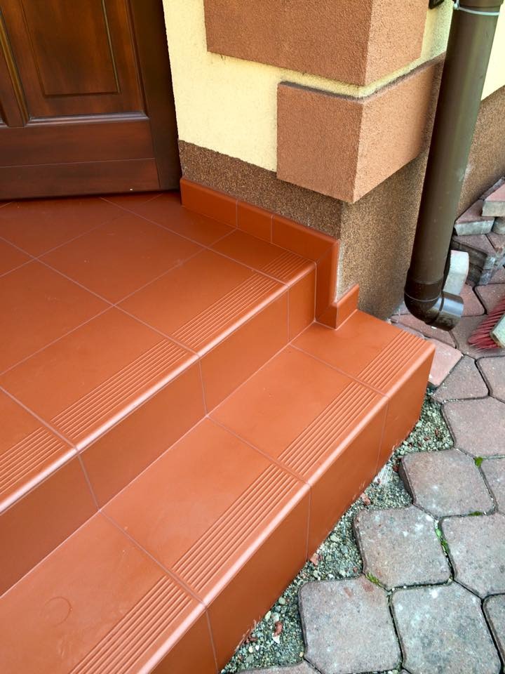 Wykończone płytkami schody wejściowe w kolorze terakoty, z antypoślizgowymi paskami, dopasowane do elewacji budynku. Widoczne detale wykończenia i kostka brukowa.