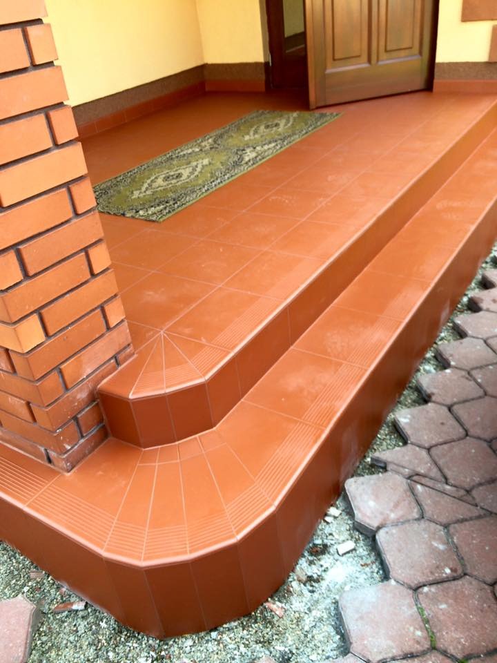 Wykończone płytkami schody wejściowe z ceglanym filarem i dywanikiem. Czerwono-brązowa kolorystyka płytek i kostki brukowej. Widoczne detale wykończenia i fuga.