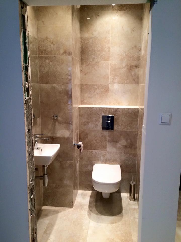 Wykończona łazienka w beżowych płytkach z podwieszaną toaletą i umywalką. Widoczne oświetlenie punktowe i fragment niedokończonego wykończenia przy futrynie.