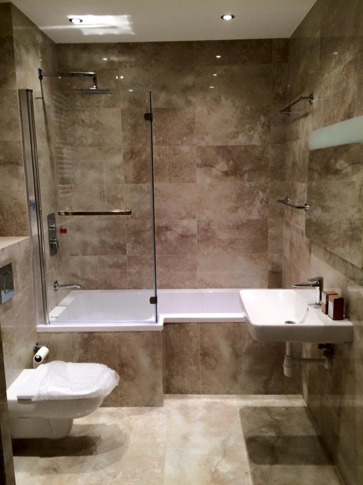 Wykończona łazienka w beżowych płytkach z wanną, kabiną prysznicową, umywalką i toaletą. Widoczne oświetlenie punktowe i akcesoria łazienkowe.