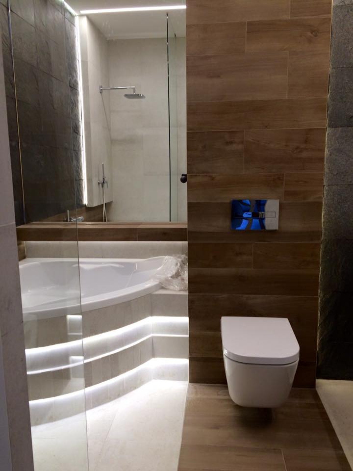 Nowoczesna łazienka z podświetlaną wanną i drewnopodobnymi płytkami. Minimalistyczna toaleta podwieszana. Szklana kabina prysznicowa z deszczownicą.