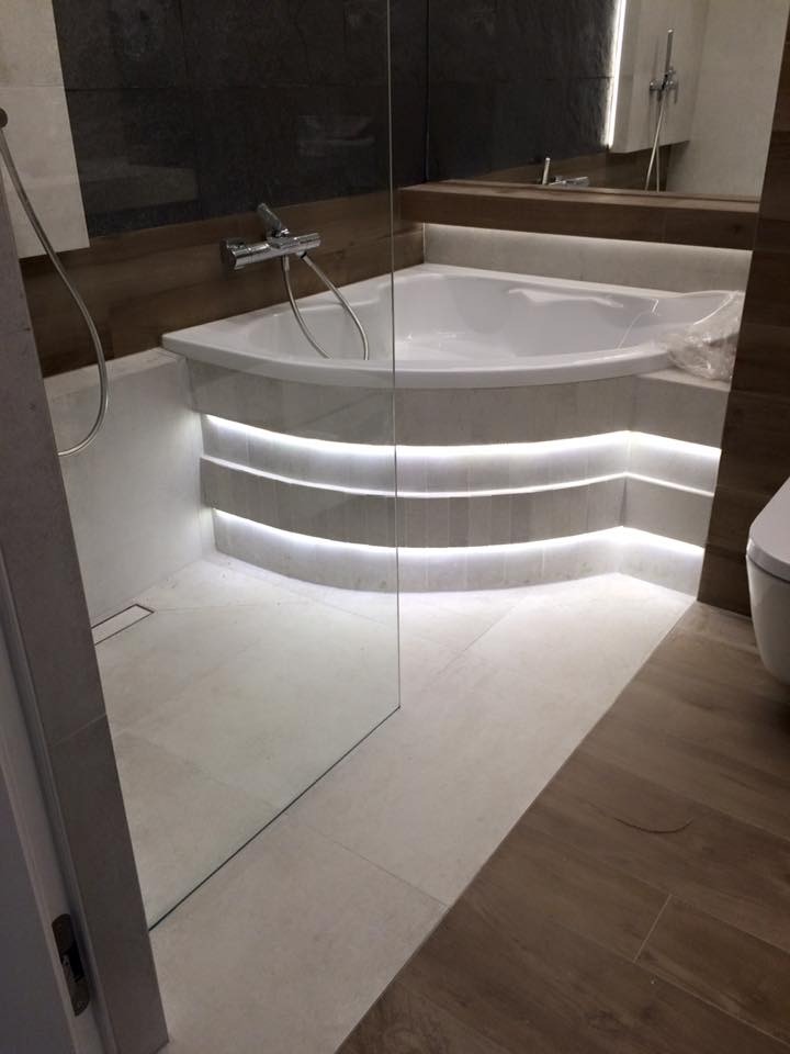 Nowoczesna łazienka z narożną wanną podświetloną taśmą LED, szklana kabina prysznicowa, płytki w odcieniach szarości i beżu, drewnopodobna podłoga.