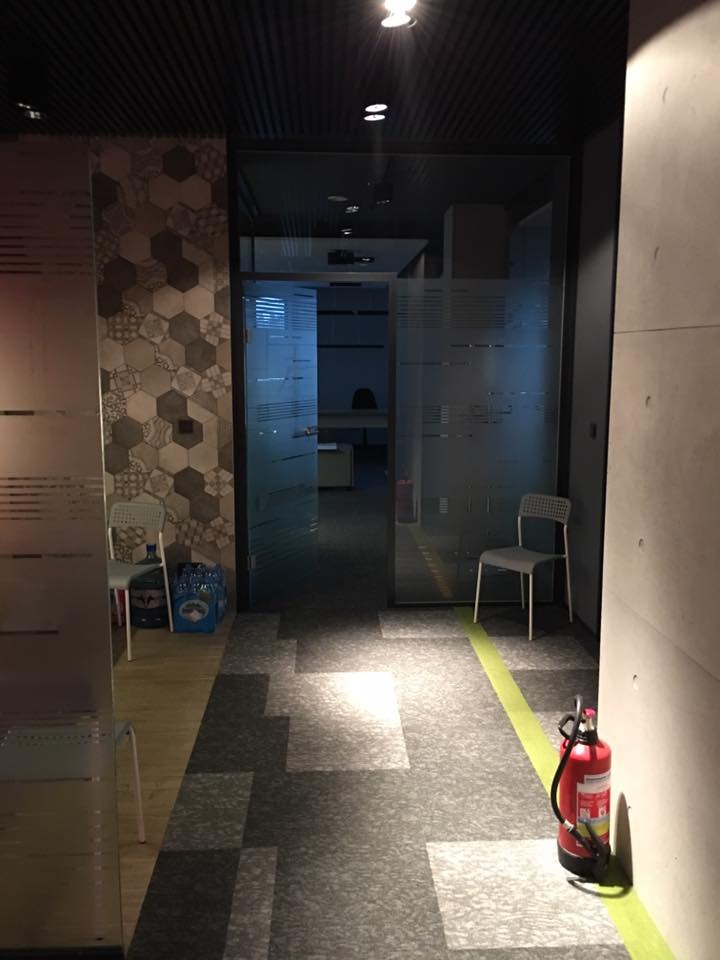 Nowoczesne biuro z przeszklonymi ścianami, wykładziną w geometryczne wzory i elementami bezpieczeństwa, takimi jak gaśnica. Widoczne krzesła i dekoracyjna ściana.