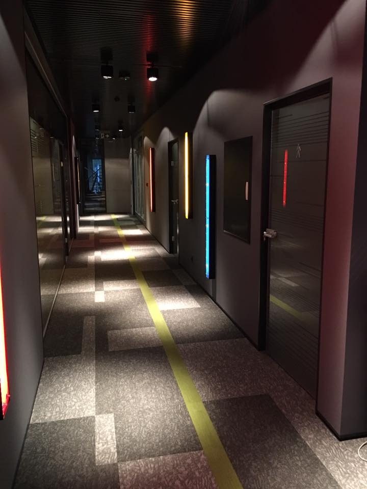 Długi korytarz biurowy z nowoczesnym oświetleniem LED w kolorach czerwonym, żółtym i niebieskim. Na podłodze wzorzysta wykładzina z żółtym pasem. Ciemne ściany i sufit.