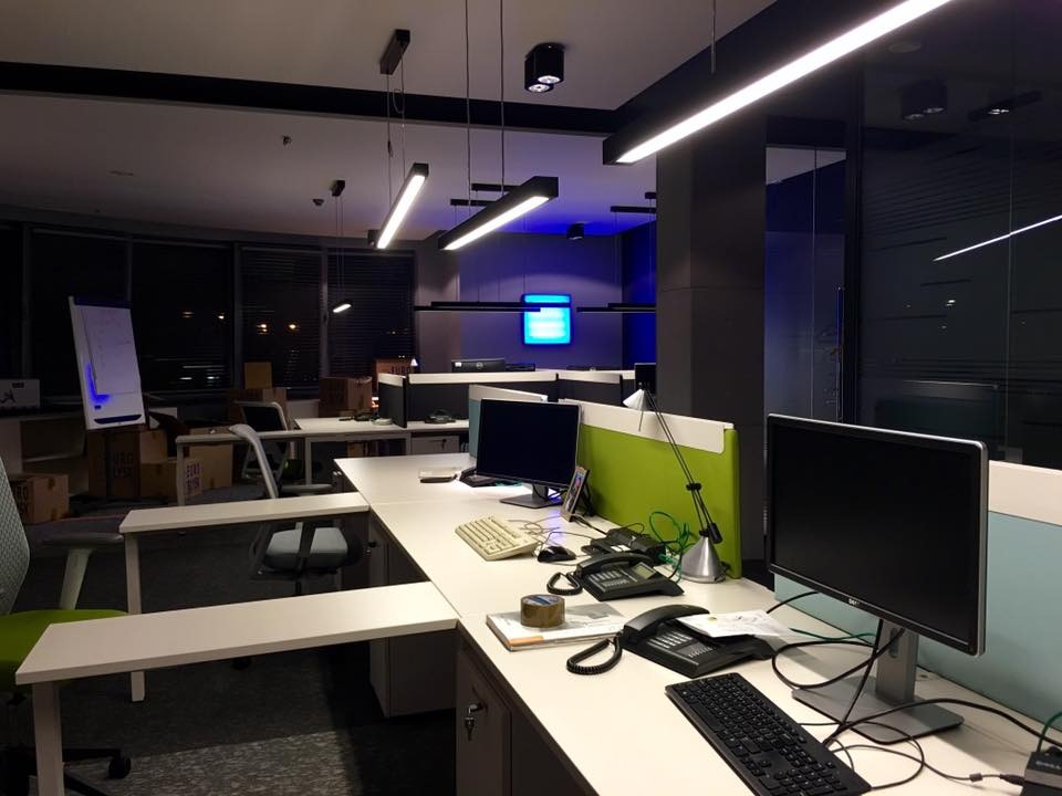 Nowoczesne biuro z podwieszanymi lampami LED, biurkami i komputerami. Widoczne elementy wyposażenia biurowego i okna. Oświetlenie sufitowe w chłodnej tonacji.