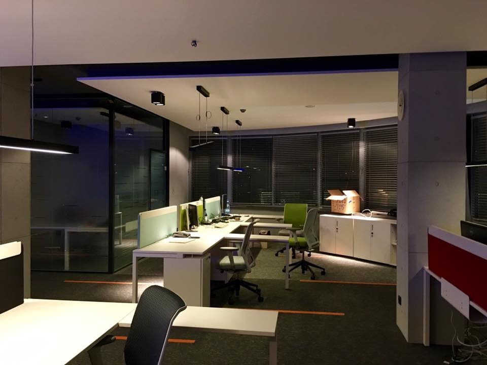 Nowoczesne biuro z oświetleniem liniowym i punktowym. Biurka z przegrodami, szare ściany, rolety w oknach. Widoczne lampy wiszące i sufitowe.