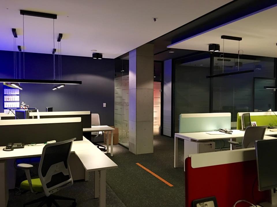 Nowoczesne biuro z minimalistycznym oświetleniem: wiszące lampy liniowe i punktowe reflektory, biurka z przegrodami w kolorach zieleni i czerwieni, szara wykładzina.