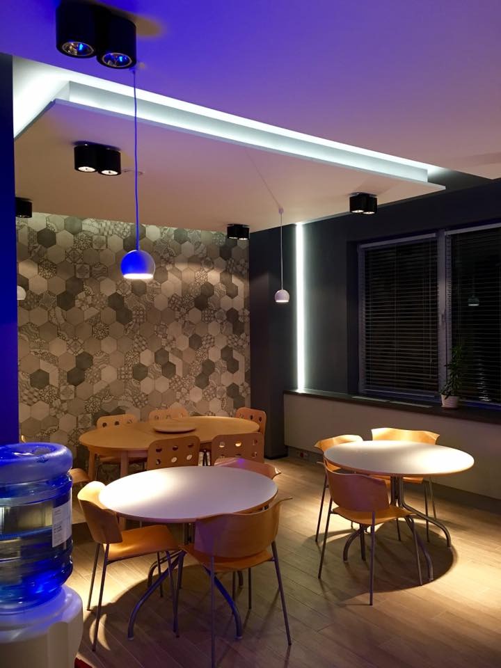 Nowoczesne oświetlenie biurowe: czarne reflektory sufitowe, podwieszana lampa z niebieską kulą i listwa LED w suficie podwieszanym. Dekoracyjna ściana z heksagonów.