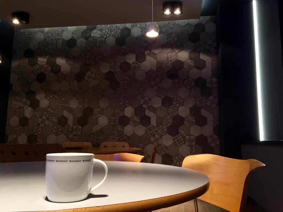 Nowoczesne wnętrze z heksagonalną tapetą, oświetleniem punktowym i minimalistycznym stołem z białą filiżanką. Akcent na detale wykończenia i subtelne światło.