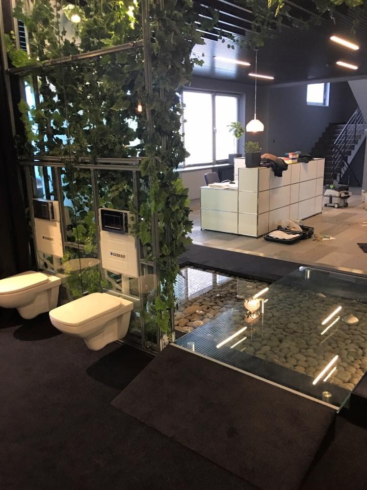 Nowoczesna aranżacja łazienki z podwieszanymi toaletami Geberit, szklaną podłogą z kamieniami i zieloną ścianą z bluszczu w przestronnym wnętrzu.