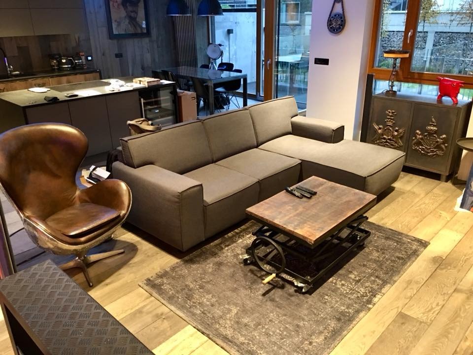 Nowoczesny salon z szarą sofą narożną, stolikiem kawowym z regulacją wysokości i skórzanym fotelem. W tle widoczna kuchnia i jadalnia, tworzące otwartą przestrzeń.