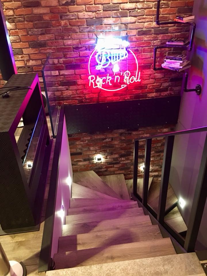 Schody z drewnianymi stopniami i czarną balustradą, oświetlone punktowymi lampami. Na ścianie neon 'Blues Rock'n'Roll' i półka z książkami. Ceglana ściana w tle.
